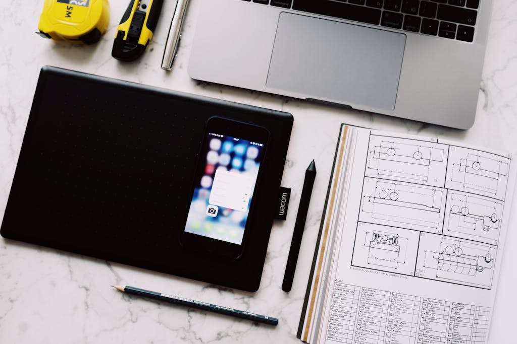 From above view of modern workplace with laptop and tablet and smartphone placed near opened notebook with drawings and pen with pencil on white marble table
